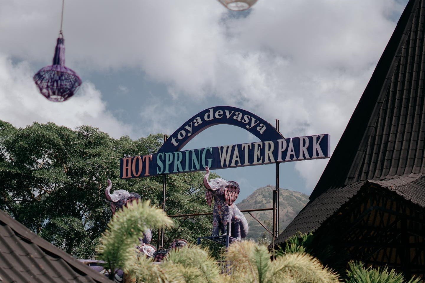 Σήμανση εισόδου για το Toya Devasya Hot Spring Water Park με διακοσμητικά αγάλματα ελεφάντων, δέντρα και βουνά στο παρασκήνιο.