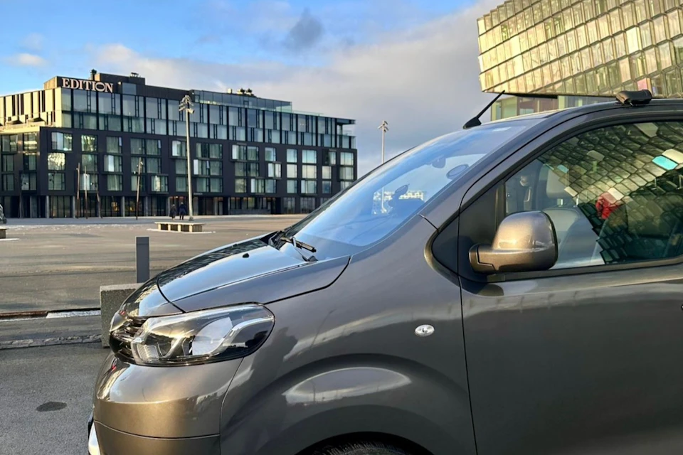Close-up of a gray van parked in front of modern glass buildings on a clear day.