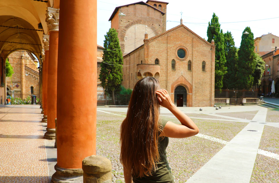 Woman with long hair standing by orange pillars, facing a historic brick church with arched windows and trees in the background.
