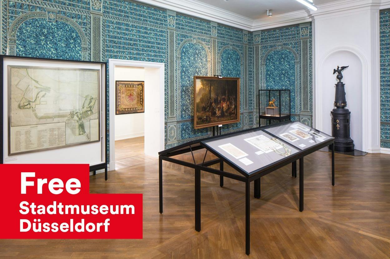Ein Ausstellungsraum in einem Museum mit kunstvoll verzierten, blau gemusterten Wänden, gerahmten Kunstwerken und gläsernen Vitrinen auf einem Holzboden.