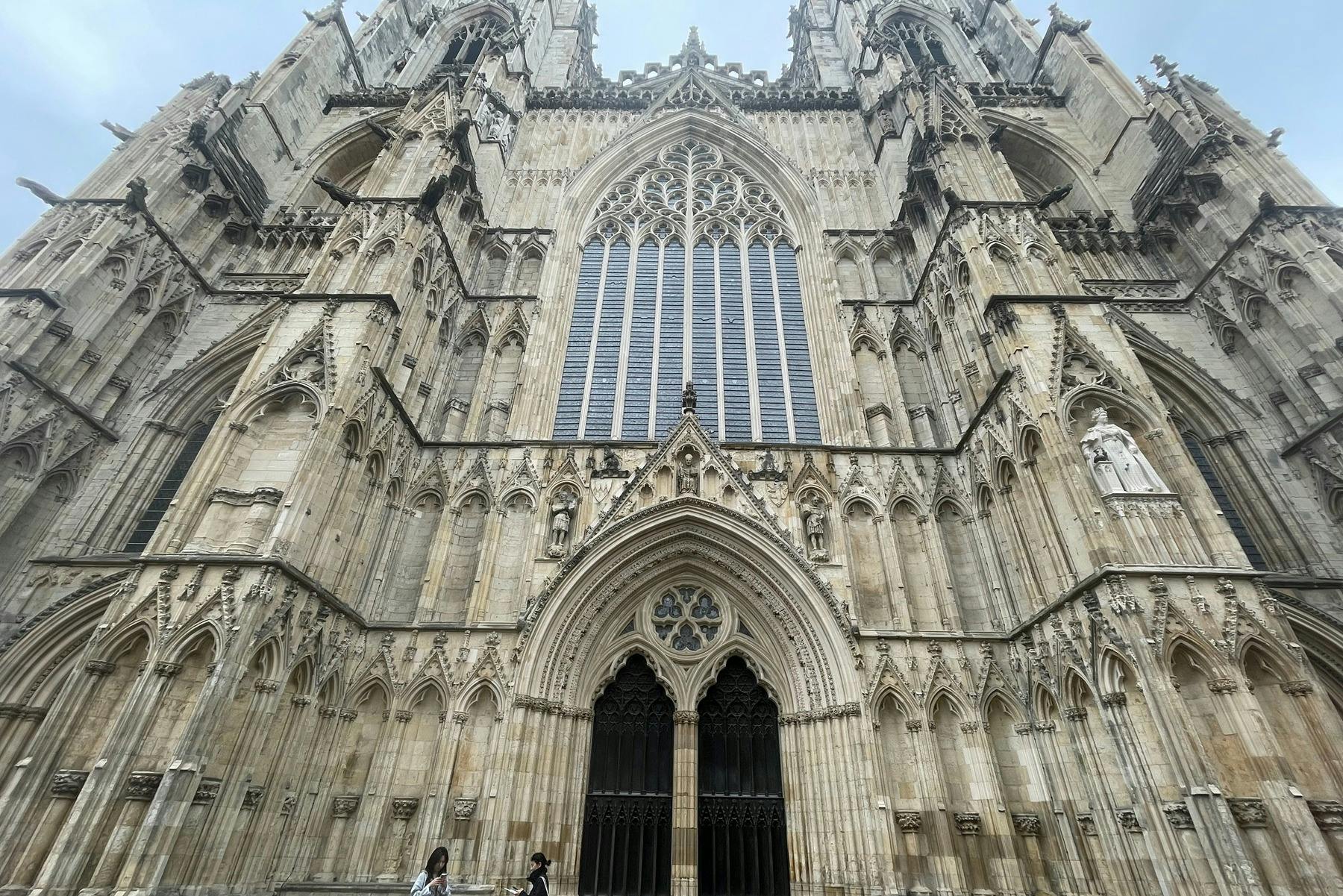 複雑な石の彫刻が施されたゴシック様式の大聖堂のファサード、2つの大きなアーチ型の入り口、背の高い中央の窓、そして下を歩く人々。