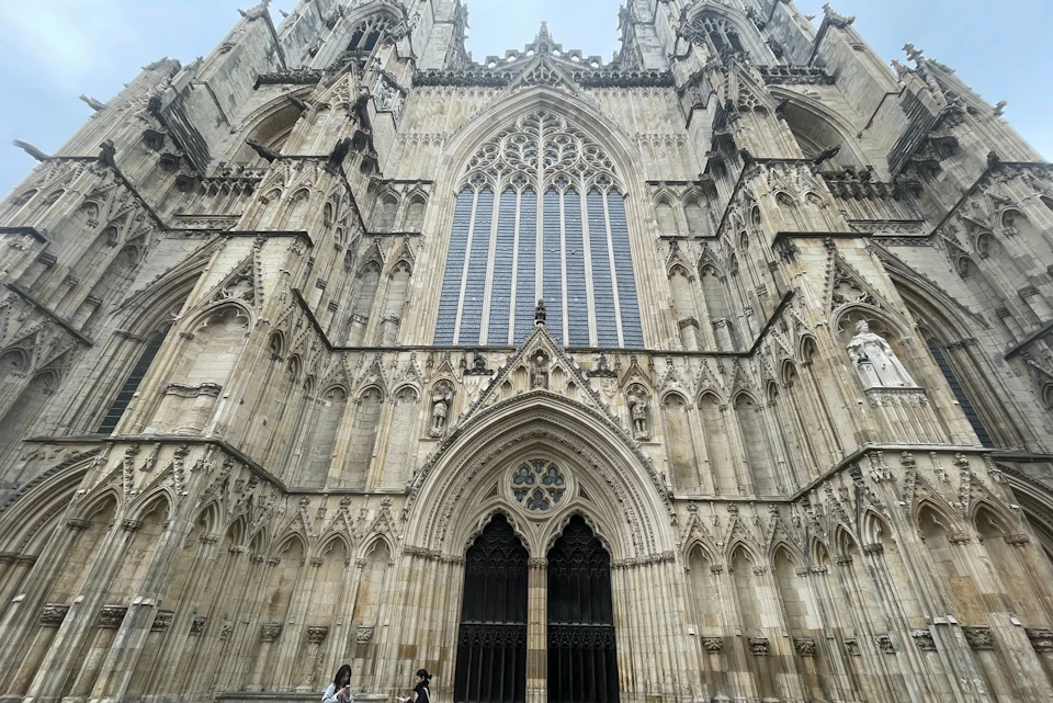 Gothic cathedral façade with intricate stone carvings, two large arched entrances, a tall central window, and people walking below.