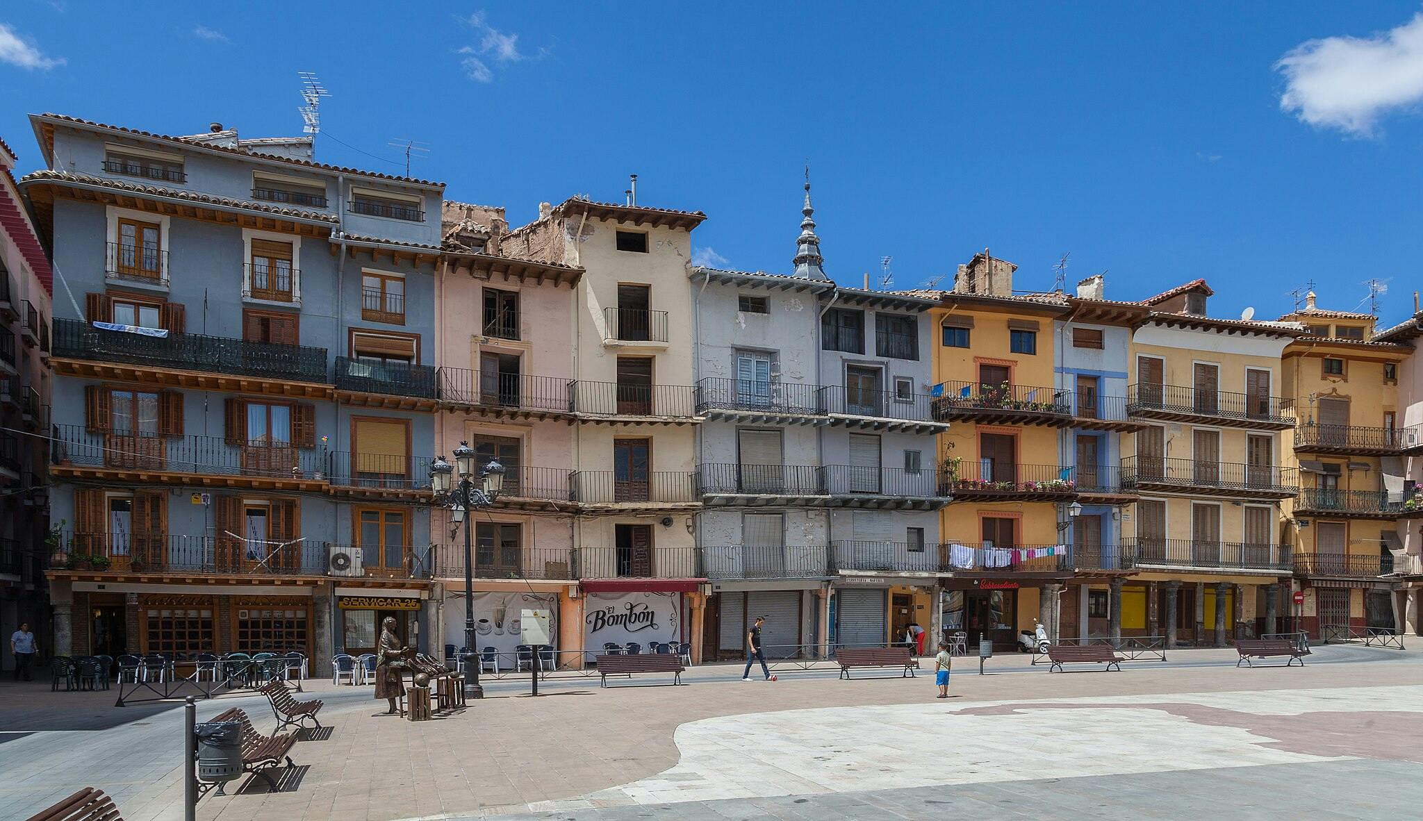 Des bâtiments colorés avec des balcons bordent une place sous un ciel bleu clair. Peu de gens se promènent et des bancs sont disséminés un peu partout.