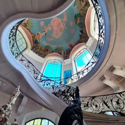 En spiraltrappa med utsmyckade svarta räcken, en takmålning och stora fönster i ett stort inomhusutrymme.