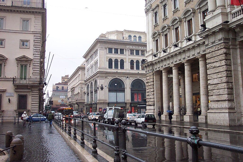 Una strada cittadina trafficata con edifici storici, marciapiedi bagnati, auto parcheggiate, un autobus e persone che camminano con gli ombrelli.