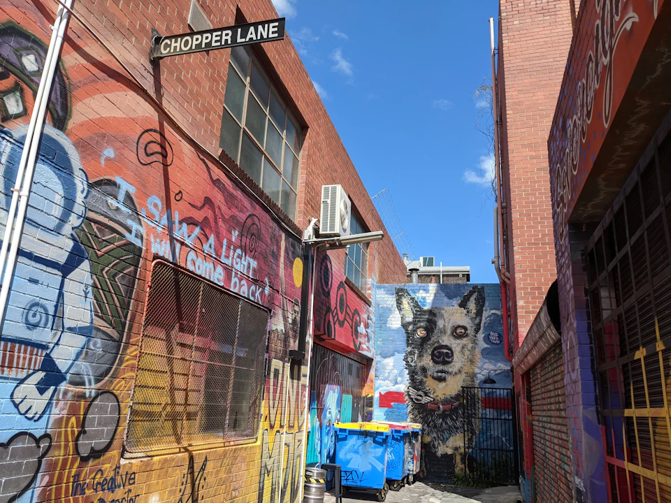 Narrow alley with colorful graffiti, a large dog mural, brick buildings, air conditioning units, dumpsters, and a "Chopper Lane" sign.