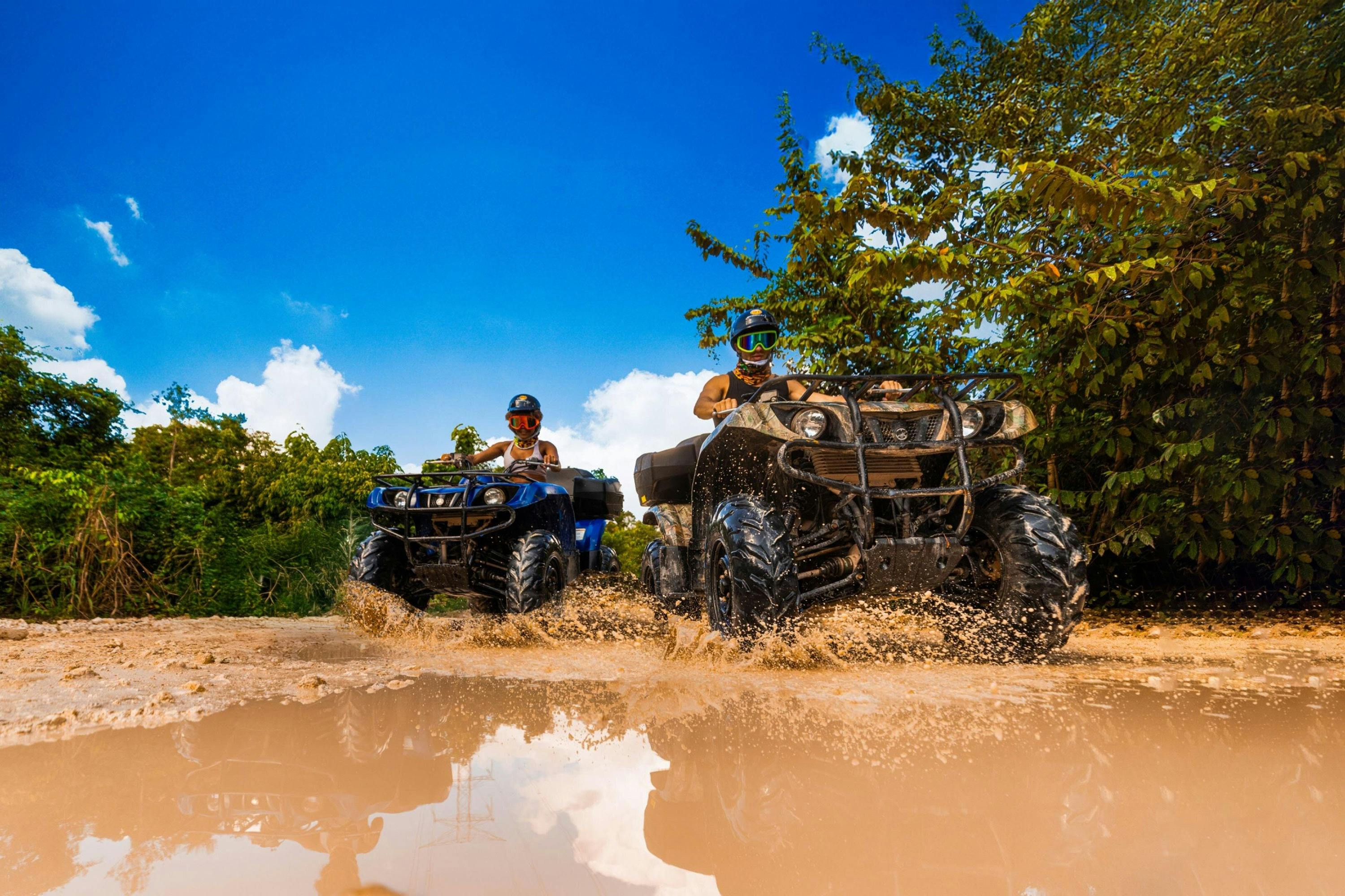 Two individuals riding all-terrain vehicles through a muddy trail surrounded by greenery under a clear blue sky.