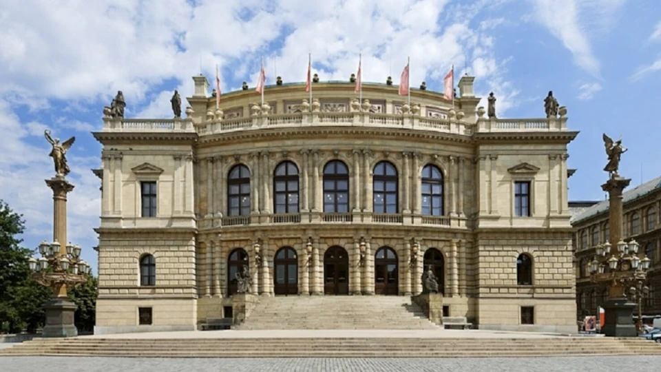 Large, ornate building with arched windows, statues, and flags on the rooftop, set against a partly cloudy sky.