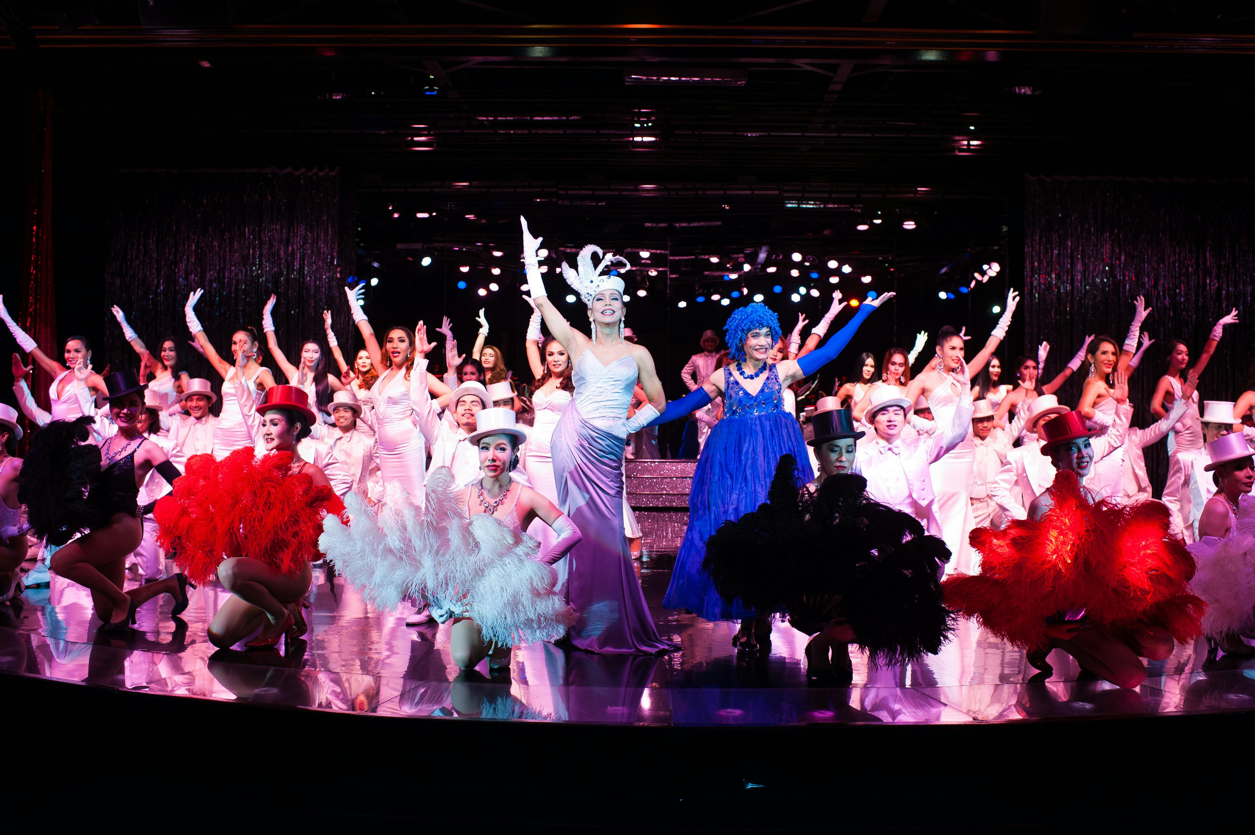 Eine große Gruppe von Tänzern in farbenfrohen Kostümen tritt auf der Bühne auf, mit hellen Lichtern und koordinierten Choreographien.