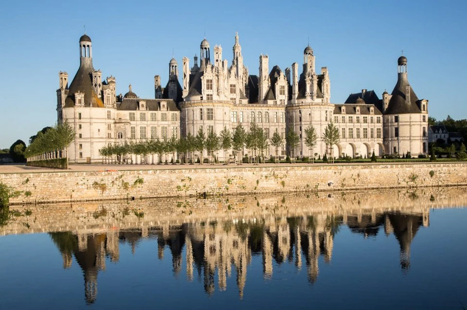 Historic château with ornate towers and turrets, reflected in a calm river under a clear blue sky.