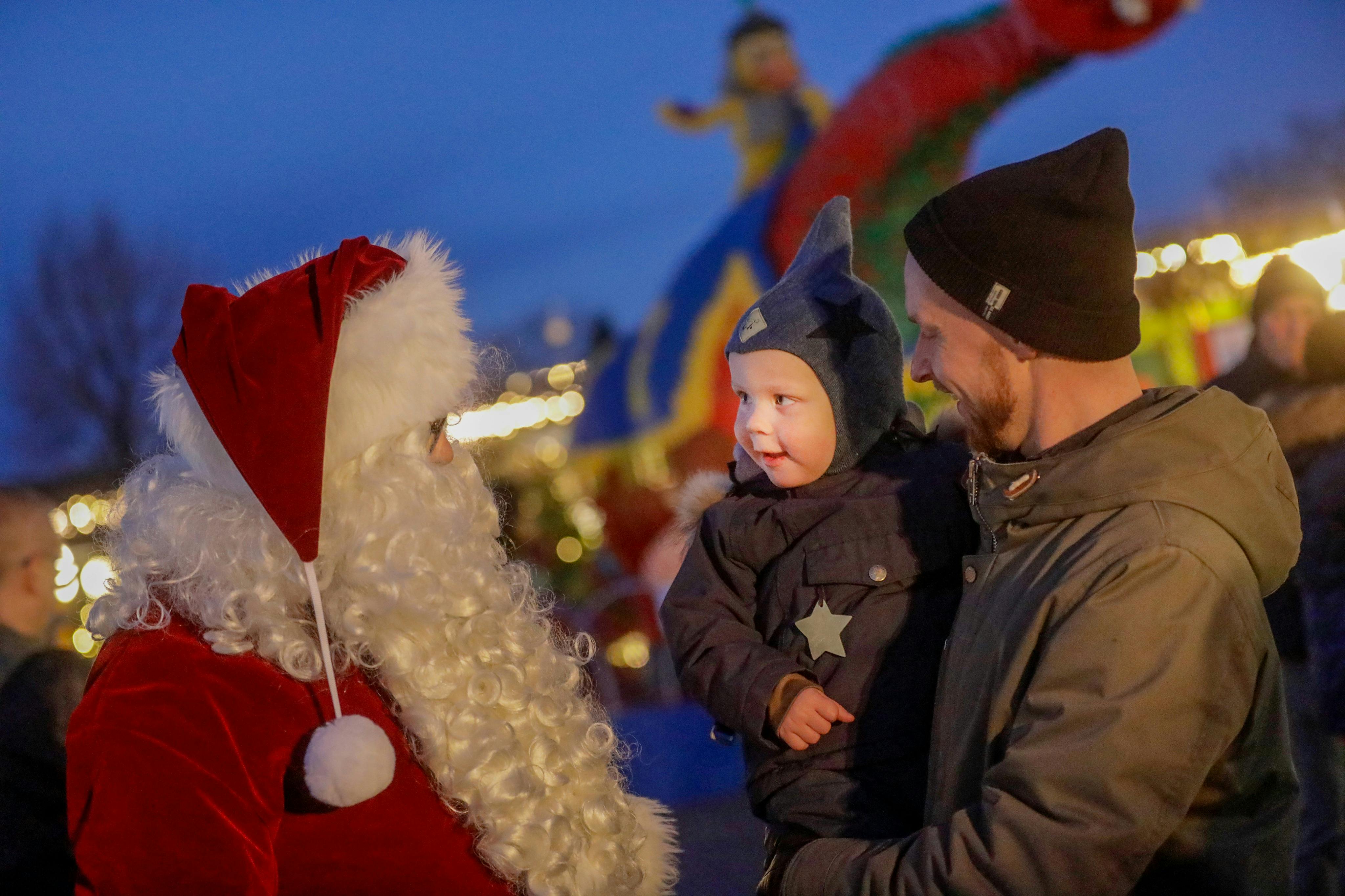 Мужчина, держащий ребенка, смотрит на человека в костюме Деда Мороза на праздничном фоне с подсвеченными украшениями.