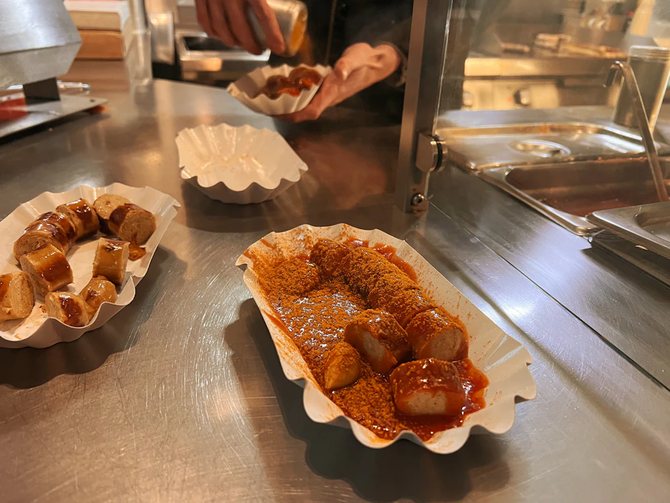 Sliced sausages covered in curry sauce and spices are served on a metal countertop in white paper trays, with a hand adding seasoning.