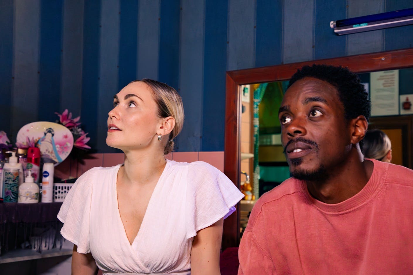 Una donna con un top bianco e un uomo con una camicia rosa guardano con attenzione fuori dalla telecamera in interni, con un muro blu sullo sfondo.