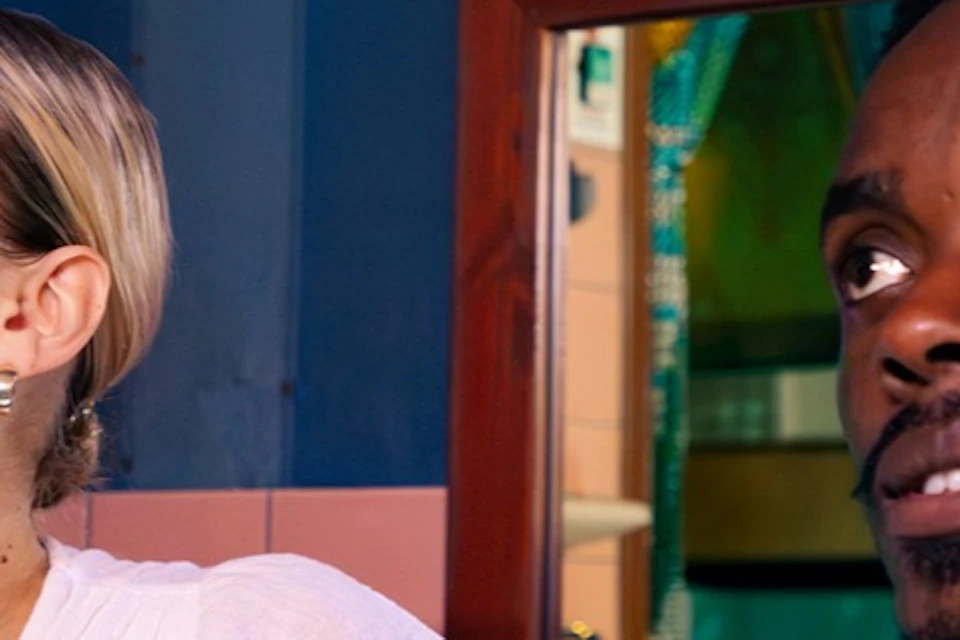 A woman in a white top and a man in a pink shirt look attentively off-camera indoors, with a blue wall in the background.
