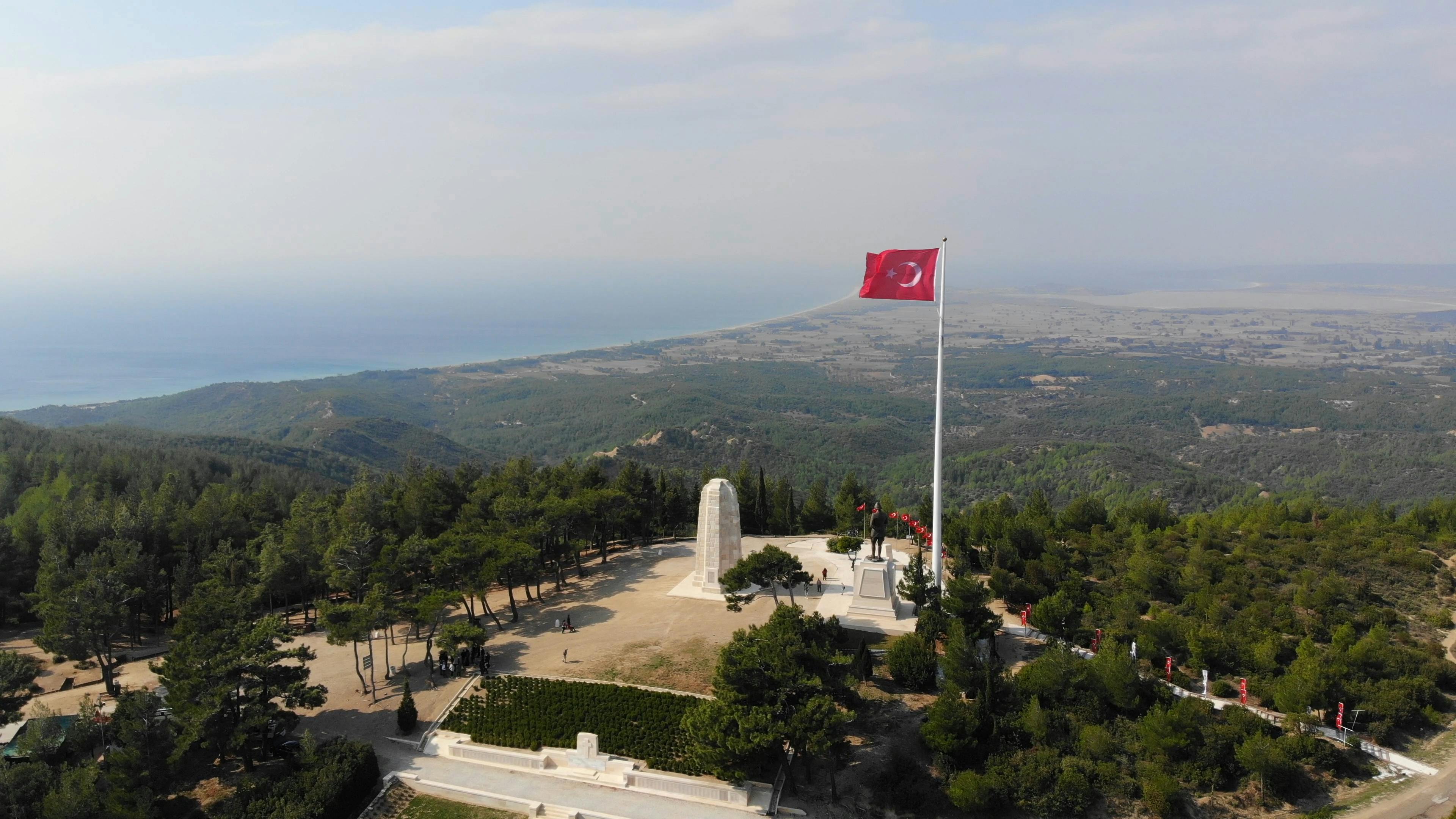Large Turkish flag flying at a hillside memorial site with surrounding trees and a panoramic view of distant landscape and sea.