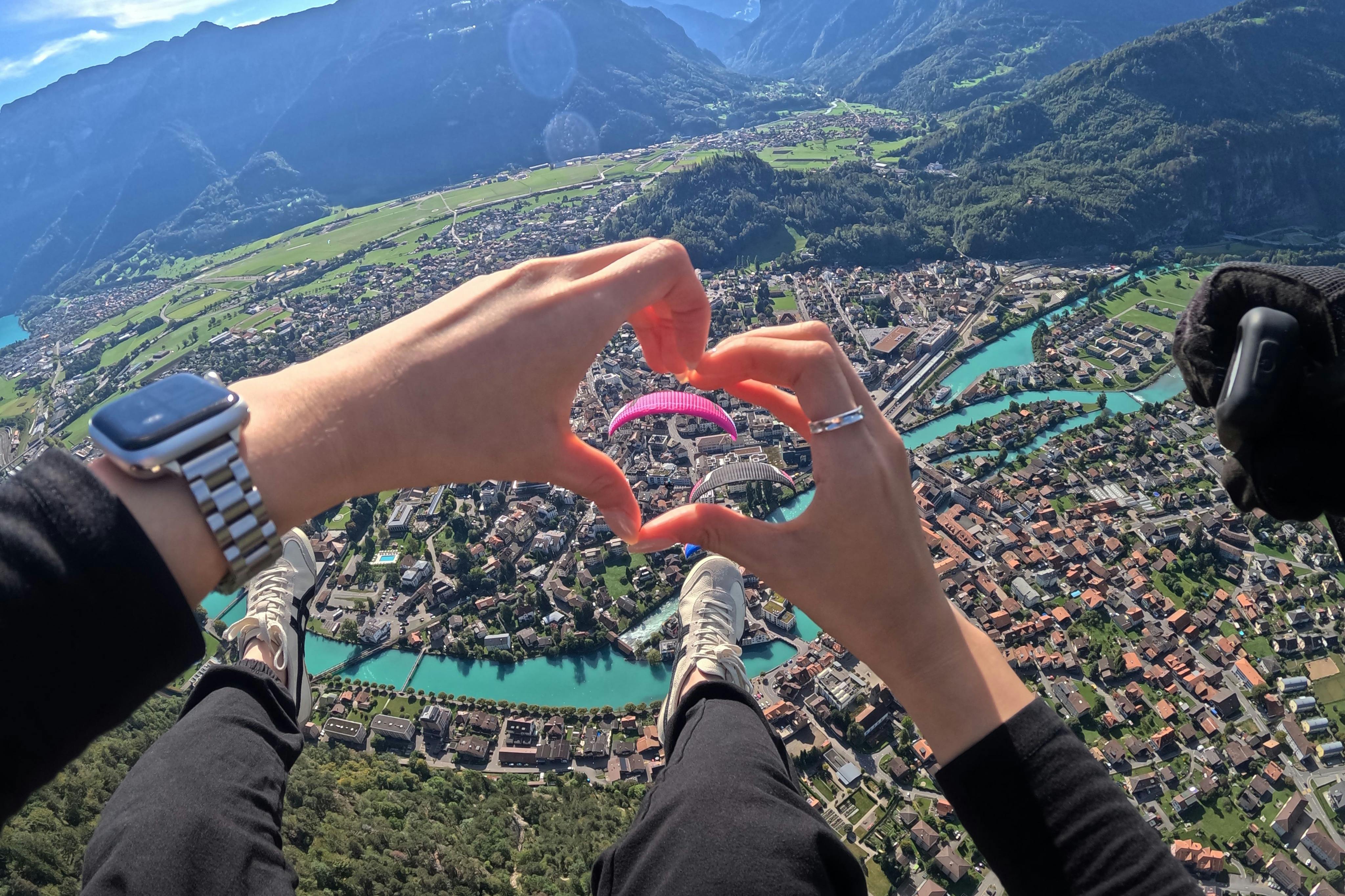 Les mains forment un cœur et les pieds sont visibles, encadrant une scène de parapente au-dessus d'une ville pittoresque avec une rivière entourée de montagnes.