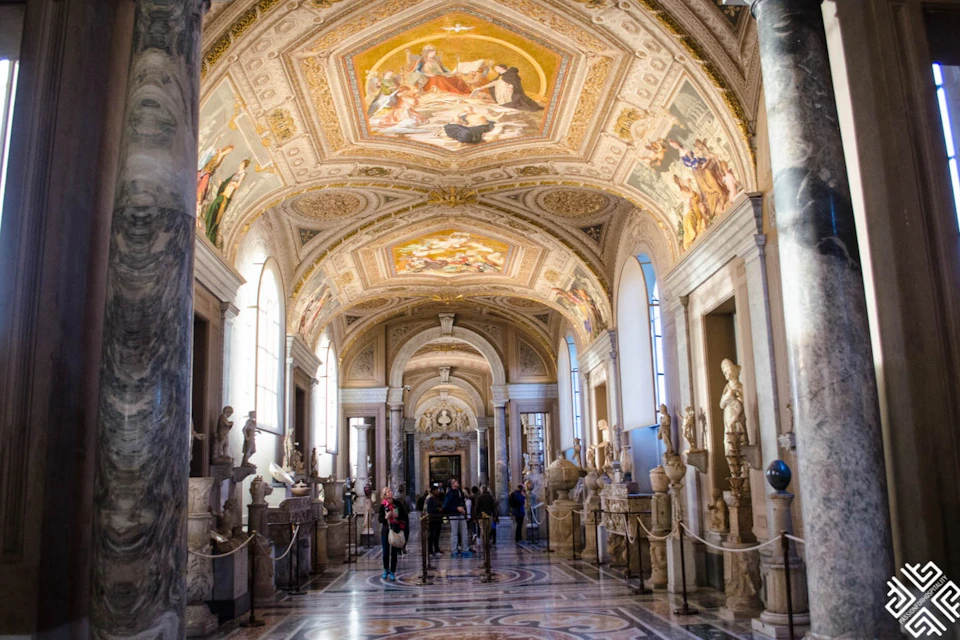A museum hallway with ornate ceilings featuring murals, arched windows, statues along the sides, and visitors walking.