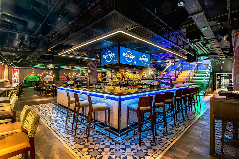 A modern bar with a square counter, stools, illuminated signs, and colorful lighting in a vibrant indoor setting.