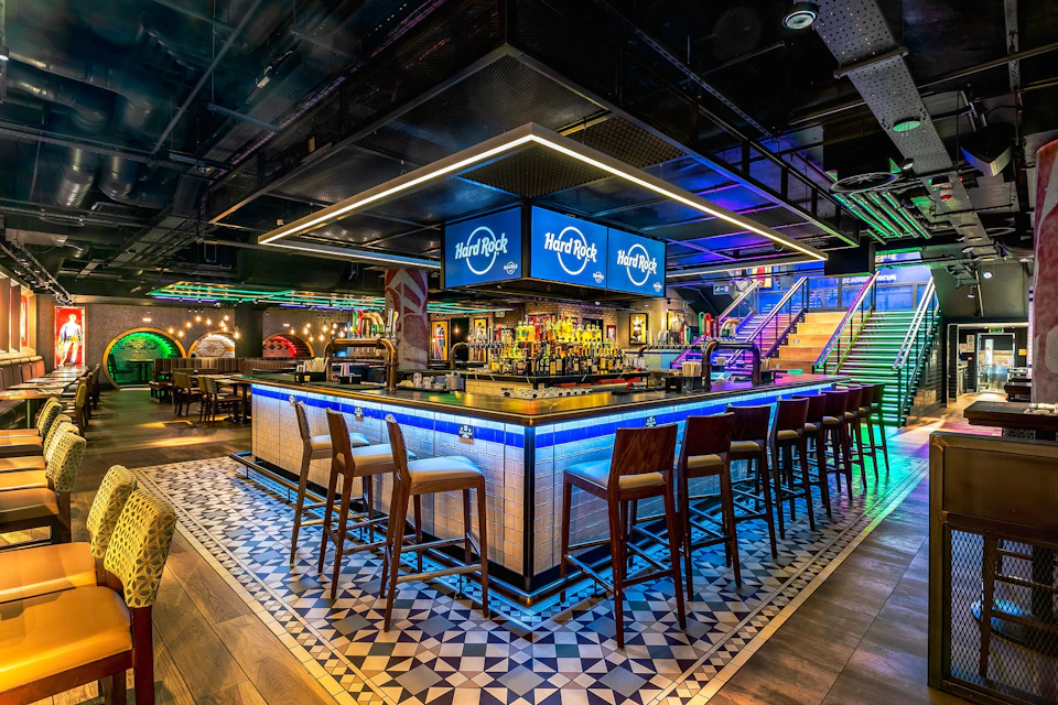 A modern bar with a square counter, stools, illuminated signs, and colorful lighting in a vibrant indoor setting.