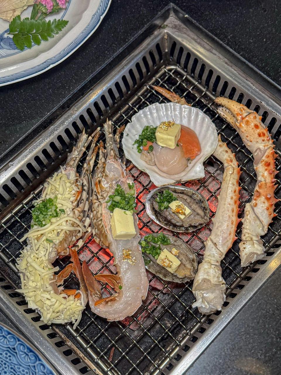 Seafood including prawns, scallops, abalone, and crab legs grilling on a metal grate over glowing coals, garnished with herbs and butter.