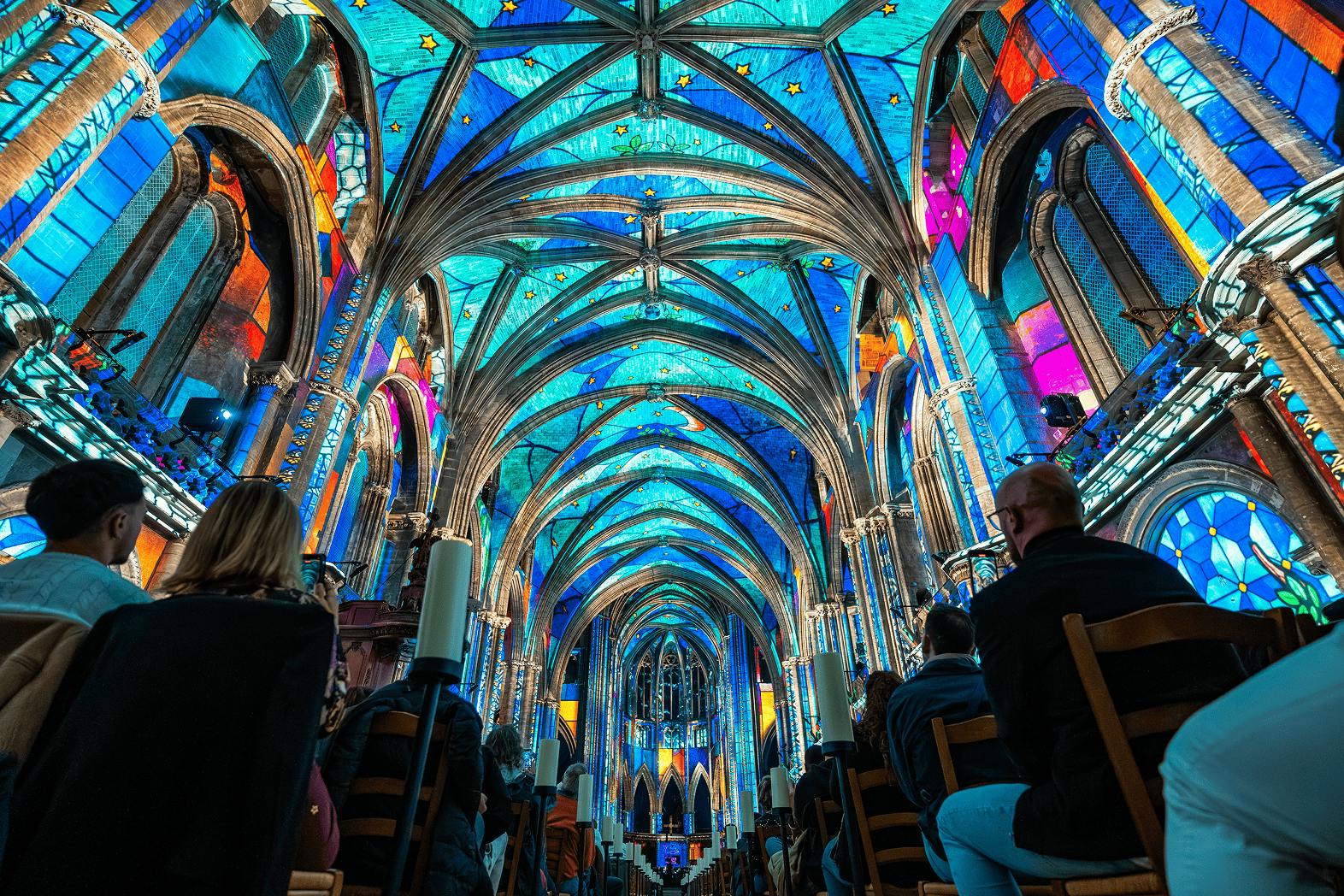 A vibrant cathedral interior with a blue stained-glass ceiling, lit candles, and people seated, looking towards the altar.