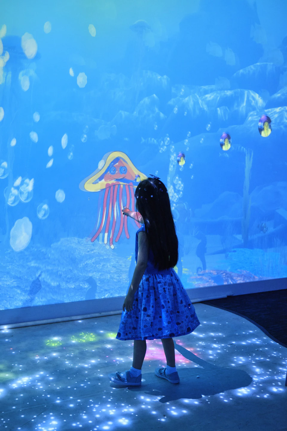 A child in a blue dress touches a glowing screen displaying an underwater scene with jellyfish and bubbles.