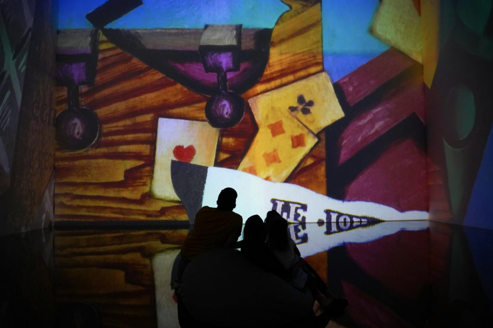 Silhouettes of three people sit in front of a colorful, abstract projection featuring playing cards and geometric shapes.