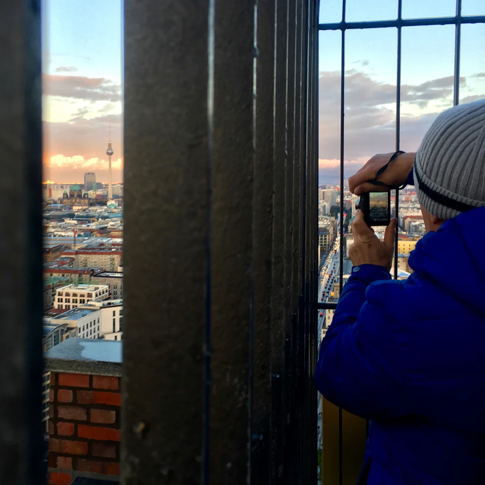 A person in a blue jacket and gray beanie takes a photo of a cityscape through metal bars on a fence or railing.