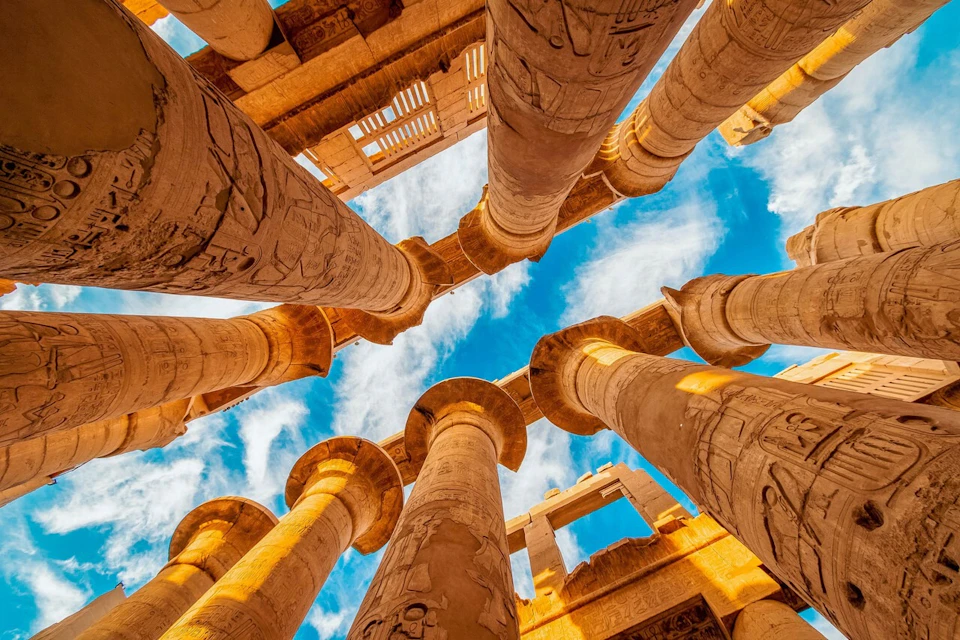 Tall ancient columns with hieroglyphs under a bright blue sky with scattered white clouds.