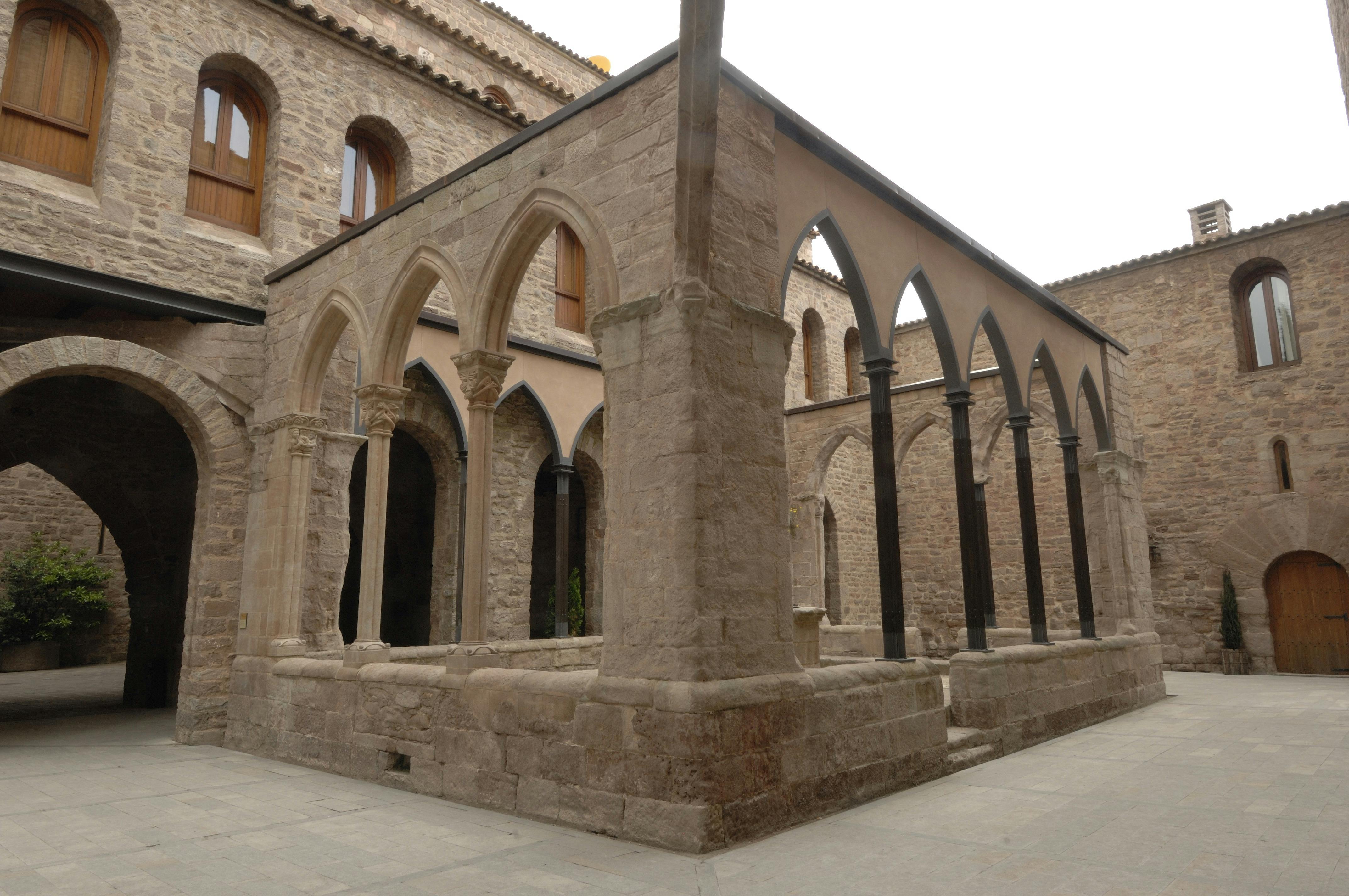石造りの中庭には、アーチ、柱、そして周囲の建物があります。 中庭の上には茶色いアーチ型の窓が見えます。