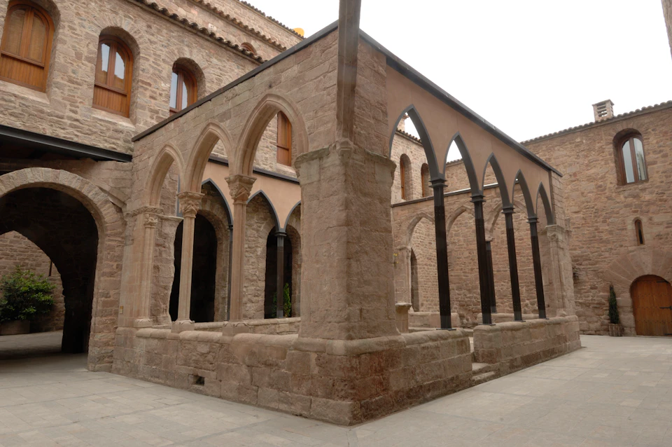 Stone courtyard with archways, pillars, and a surrounding building. Brown arched windows are visible above the courtyard.
