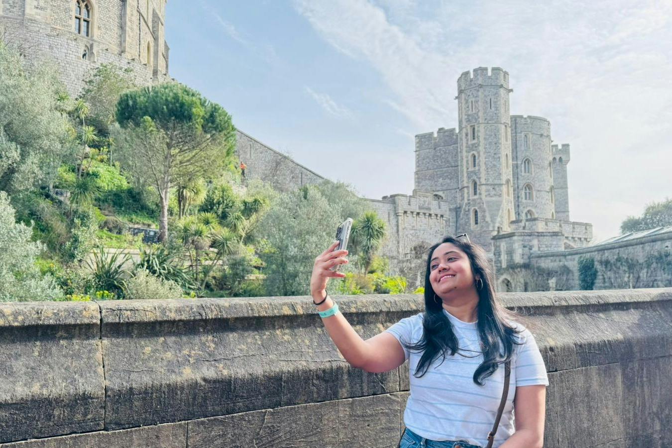 A person taking a selfie in front of a historic stone castle on a clear day, with greenery and a stone wall in the background.