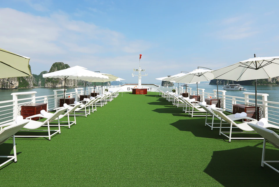 Rooftop deck of a boat with green flooring, white lounge chairs, and umbrellas. Calm water and rocky islands in the background.