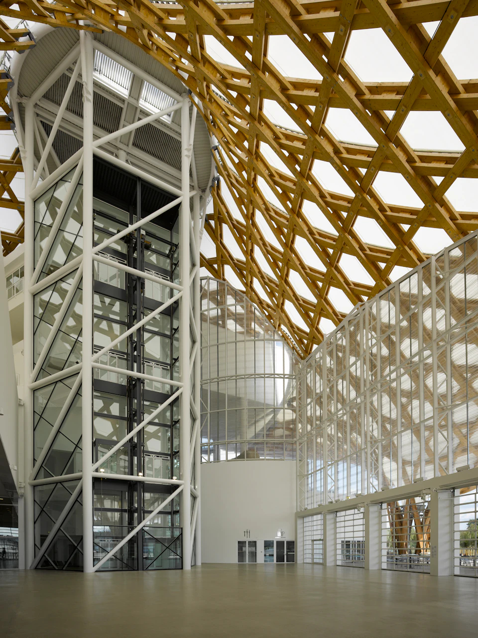 Interior of a modern building featuring a glass elevator, large glass walls, and a wooden lattice ceiling.
