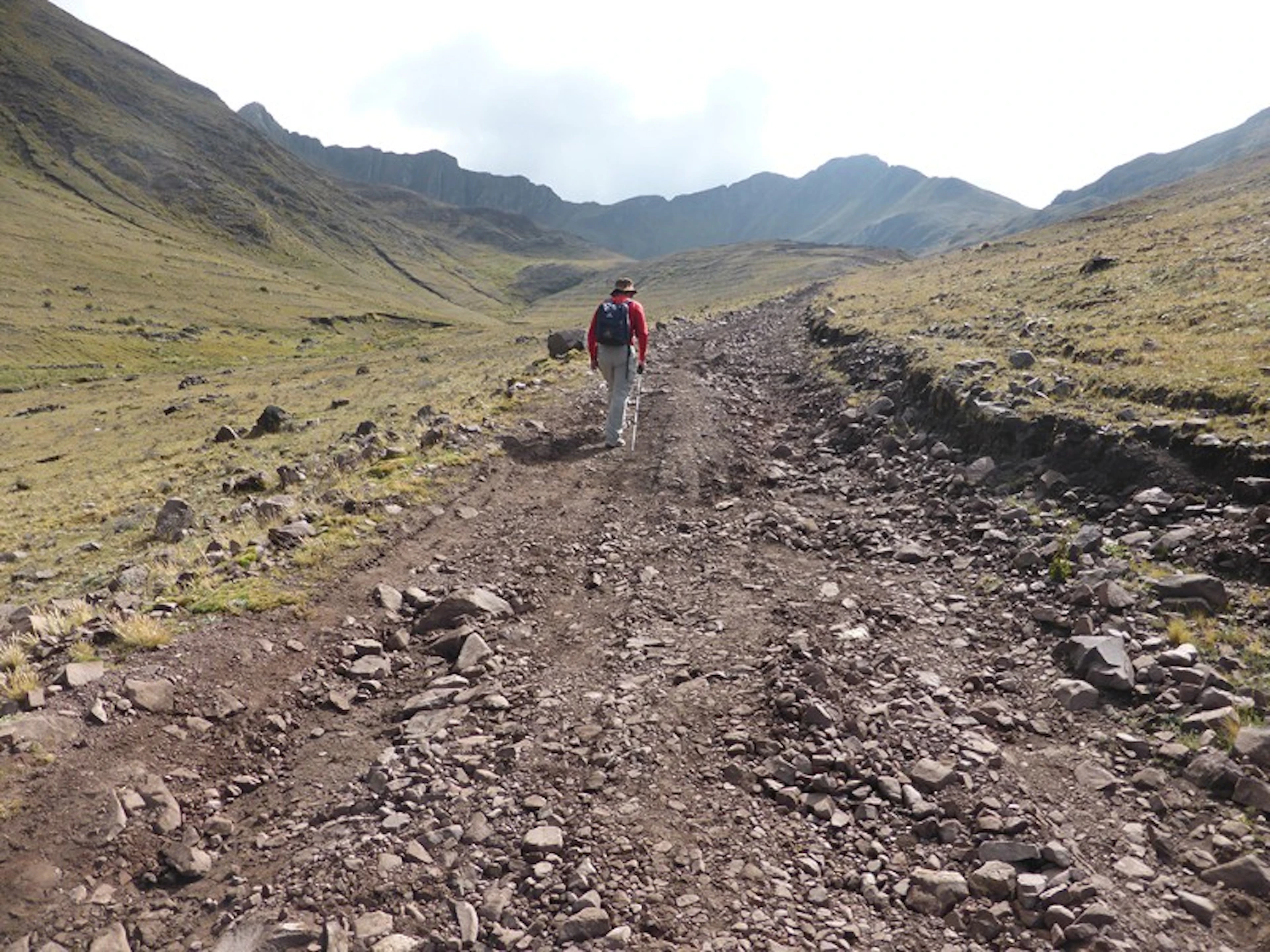 Person hiking on a rocky dirt trail surrounded by grassy hills and mountains under a cloudy sky.
