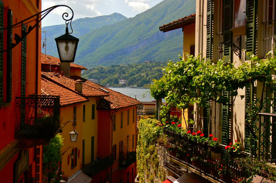 Narrow alley with colorful buildings and balconies filled with flowers, overlooking a lake with mountains in the background.