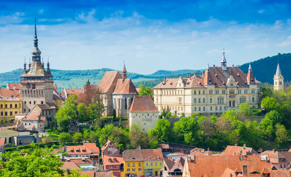 Historic buildings with red-tiled roofs are surrounded by greenery, with hills under a vibrant blue sky in the background.