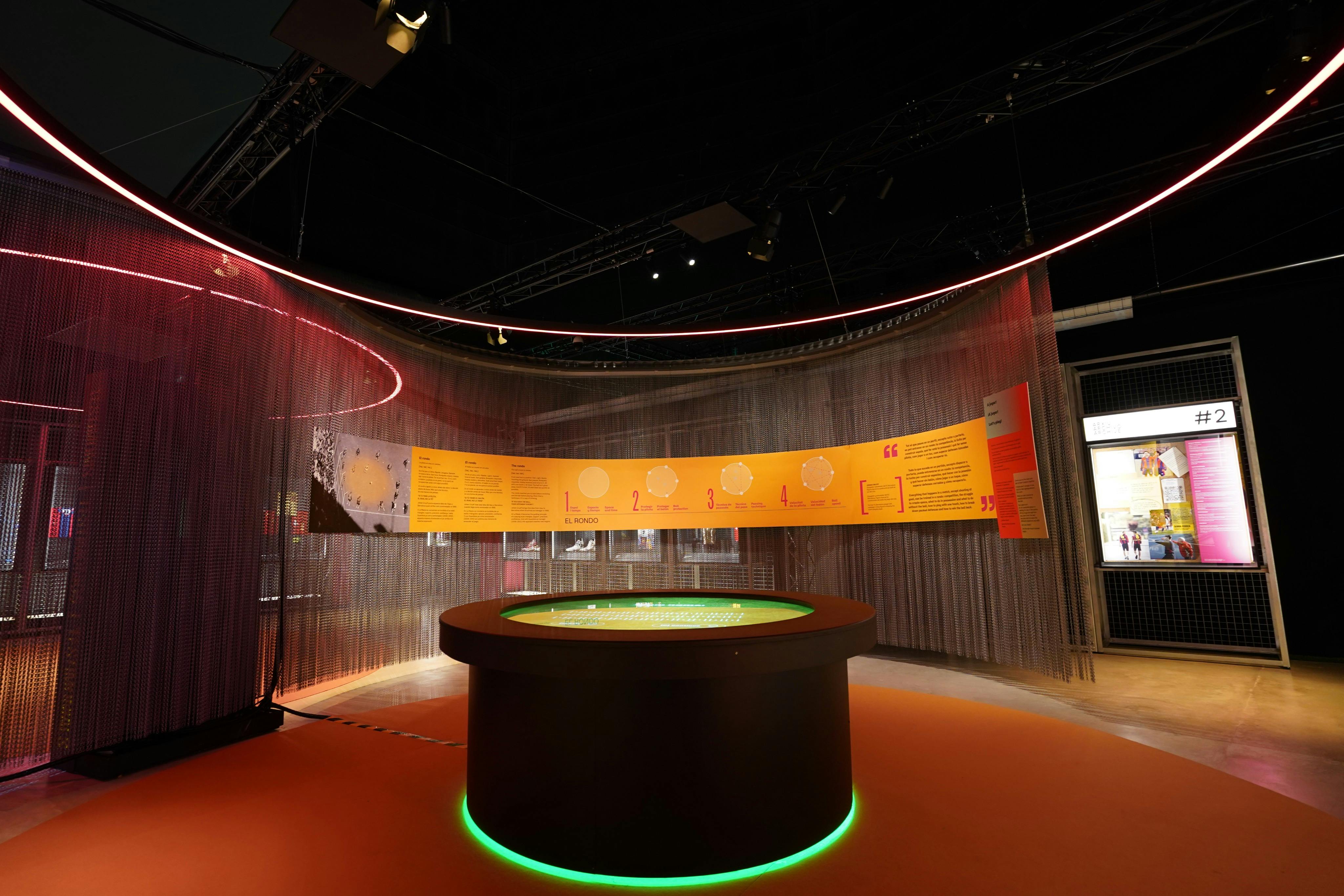 A modern exhibition setup with a circular table under green lighting and informational panels on a curved wall.