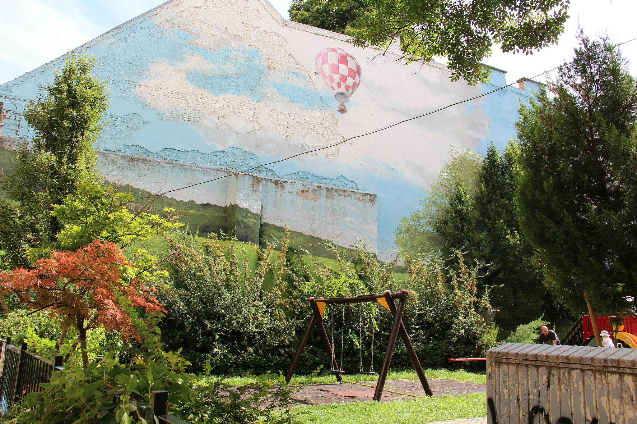 Urban väggmålning av landsbygden med luftballong, grönska, en lekplatsgunga och två barn i hörnet.