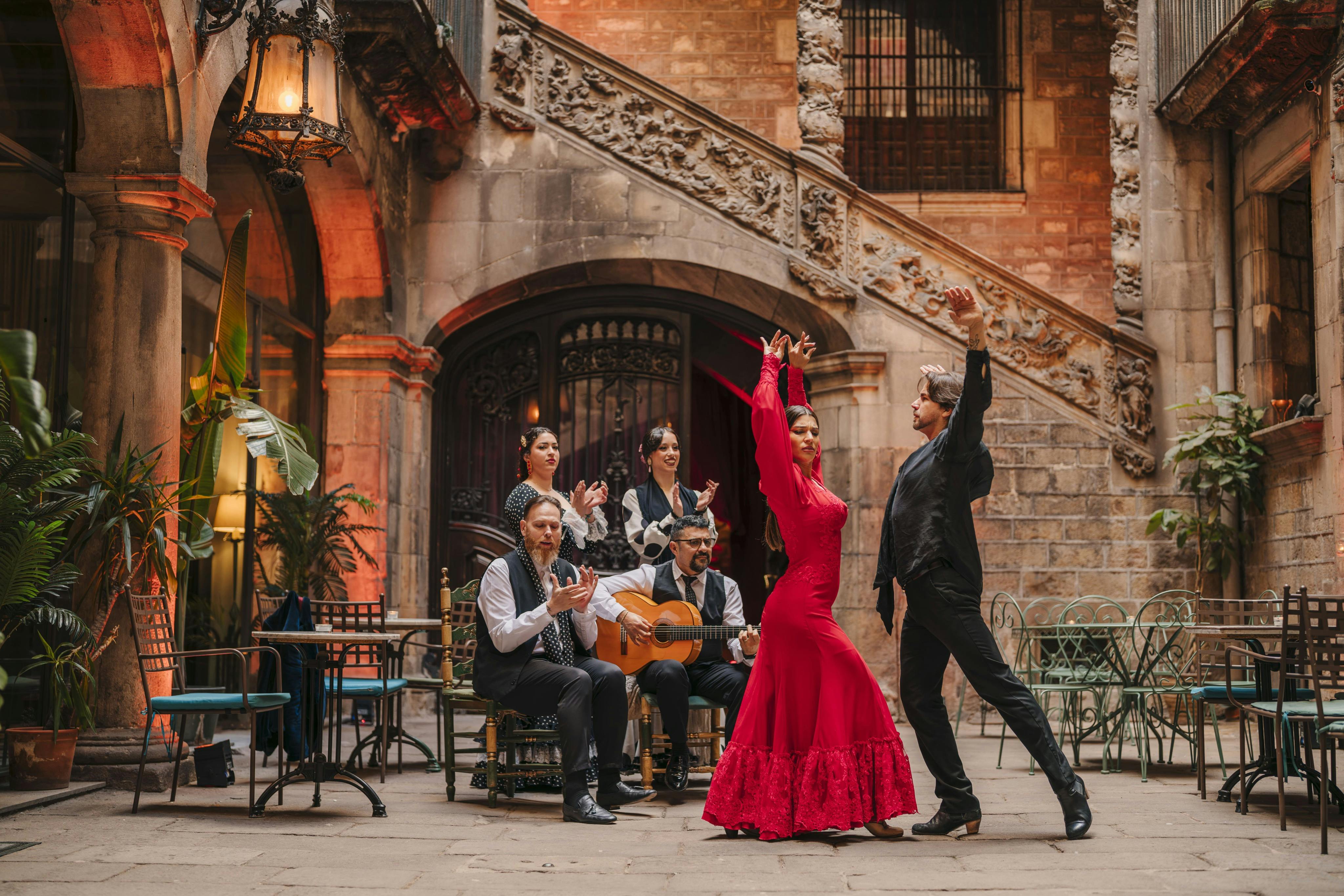 Two dancers performing a flamenco dance in a courtyard with musicians seated behind them, playing guitar and clapping.