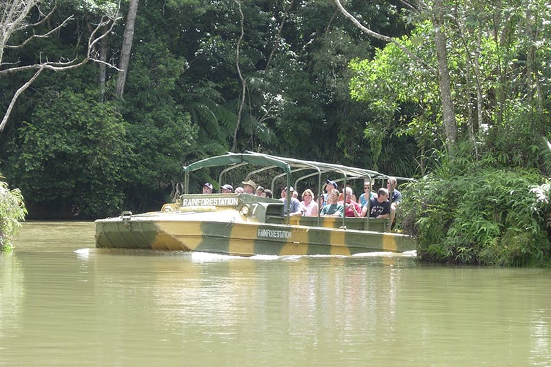 Tourists in an amphibious vehicle ride through a jungle waterway labeled "Rainforeststation" surrounded by lush greenery.