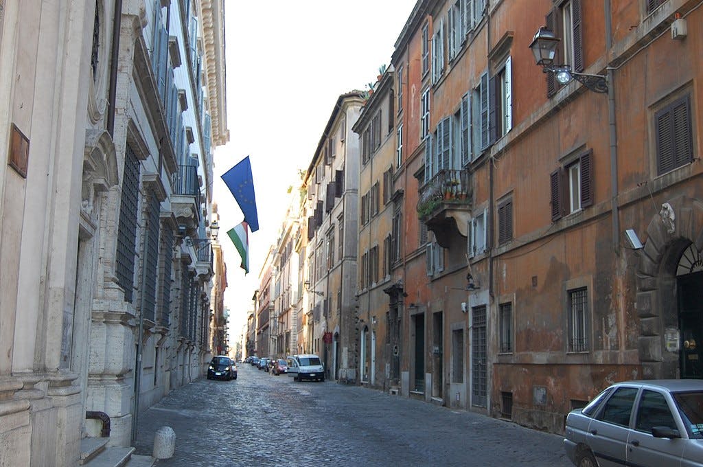 Una stretta strada di ciottoli fiancheggiata da alti edifici con bandiere europee, auto parcheggiate e finestre chiuse.