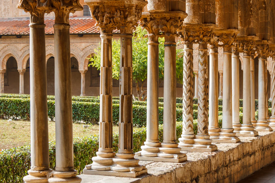 Ornate stone columns with intricate carvings line a corridor, overlooking a lush garden with neatly trimmed hedges and trees.