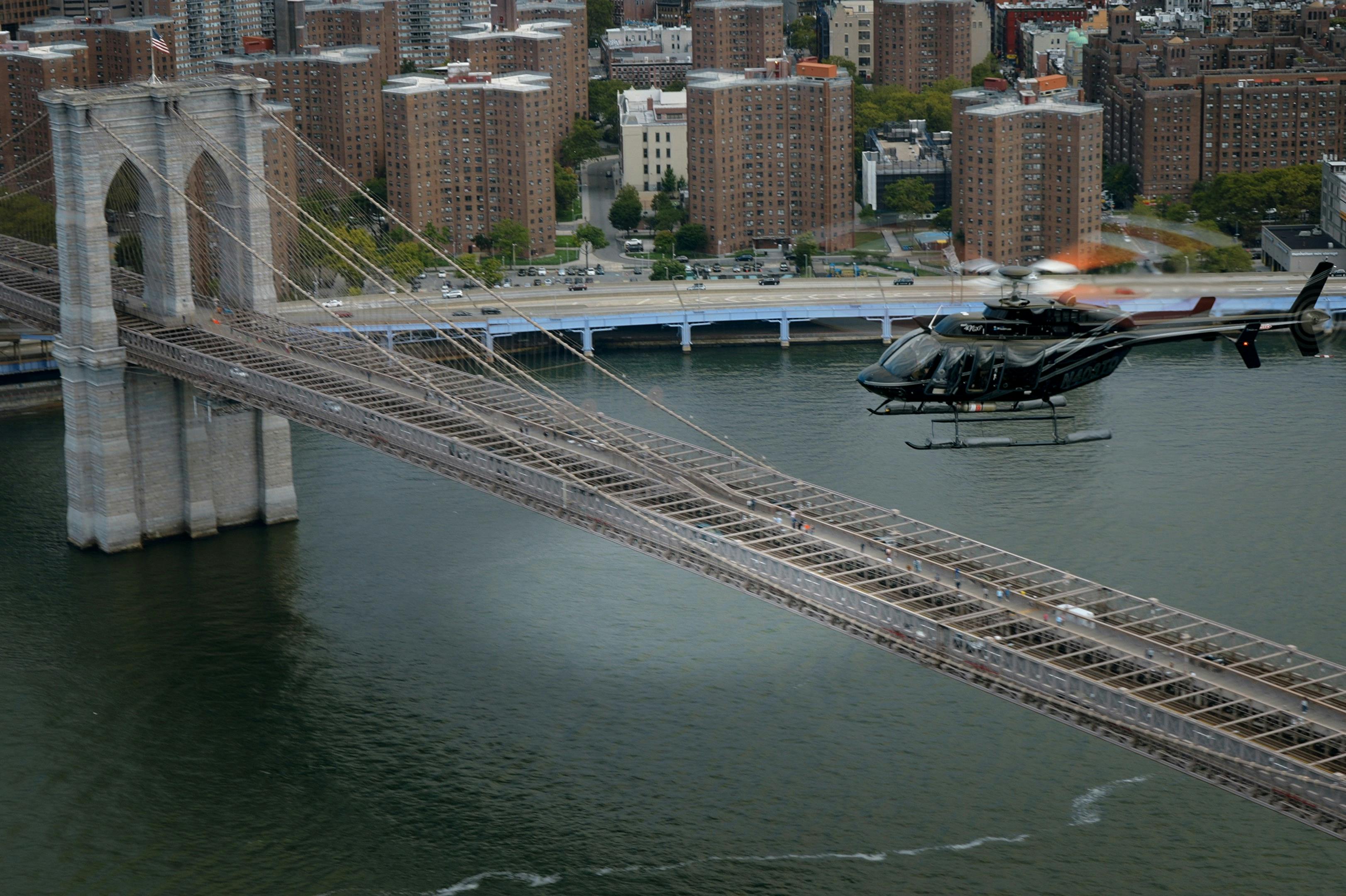 Um helicóptero a voar perto de uma ponte sobre um rio, com edifícios da cidade e uma estrada com carros ao fundo.