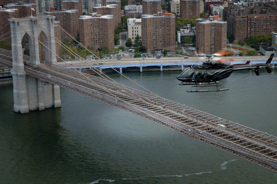 A helicopter flying near a bridge over a river, with city buildings and a roadway with cars in the background.
