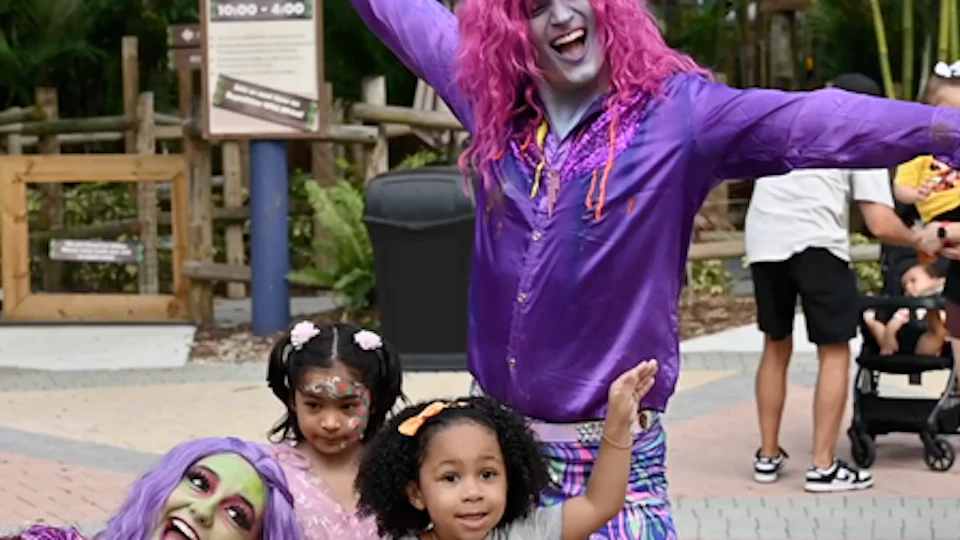 A group of performers with colorful costumes posing with children in a theme park setting, surrounded by trees and a sign.