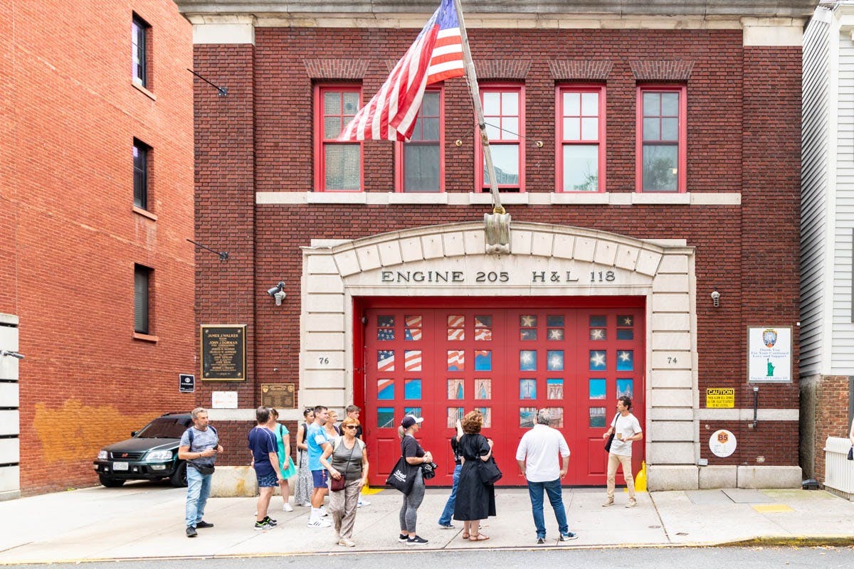 People standing in front of a fire station with red doors and an American flag flying above. The building is labeled "ENGINE 205 H&L 118."