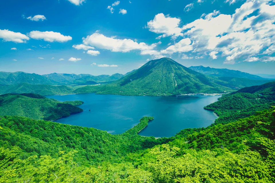 A scenic lake surrounded by lush green hills and mountains under a bright blue sky with scattered clouds.