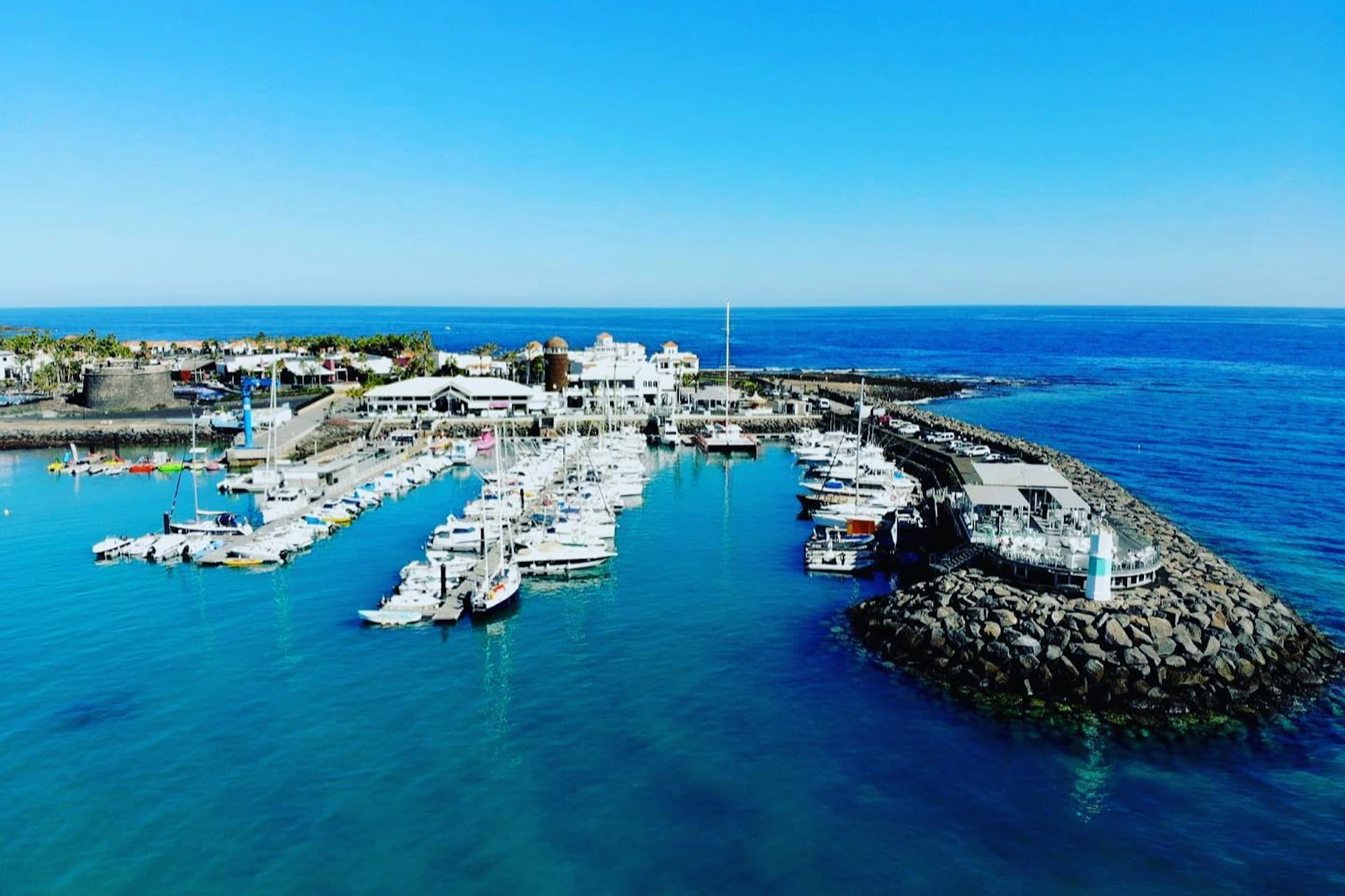 A marina with numerous docked boats, surrounded by buildings and breakwaters, set against a clear blue sky and ocean.