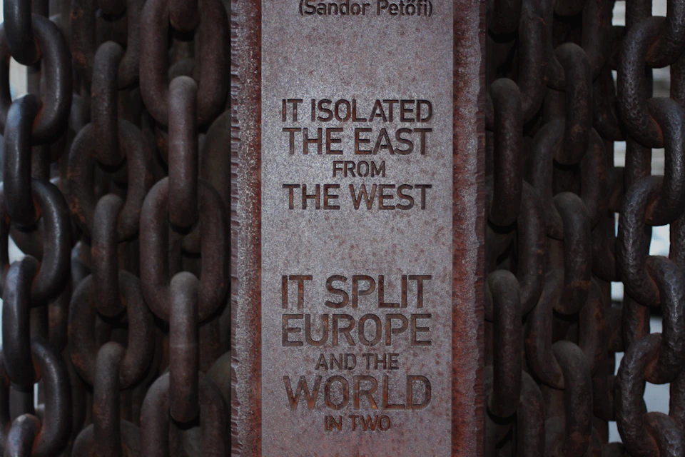 A vertical metal plaque with the text: "IT ISOLATED THE EAST FROM THE WEST. IT SPLIT EUROPE AND THE WORLD IN TWO." Chains are visible in the background.